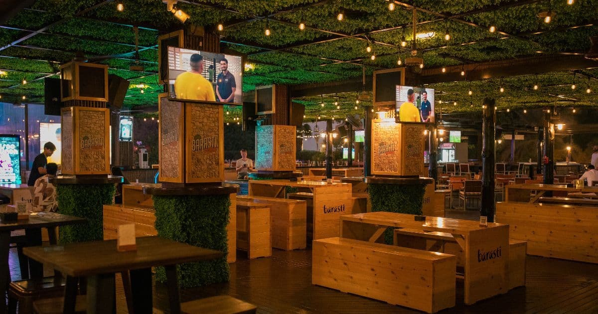 An indoor seating area at Barasti, featuring wooden benches and tables, green decorative elements on the pillars, and TV screens displaying sports, creating a cozy and vibrant atmosphere.