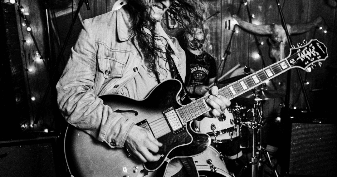 A black-and-white photo of a musician playing an electric guitar, captured mid-performance. The guitarist's long hair and denim jacket add to the rock and roll atmosphere of the scene.