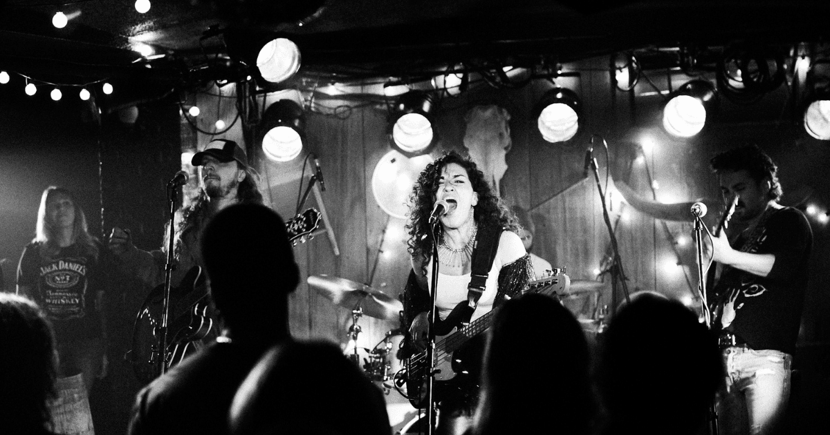 A black-and-white photo of a band performing on stage, with the lead singer passionately singing into the microphone while playing the bass guitar. The drummer and another guitarist are visible in the background, and the audience is seen watching the performance, captured in the low-light environment of the venue.