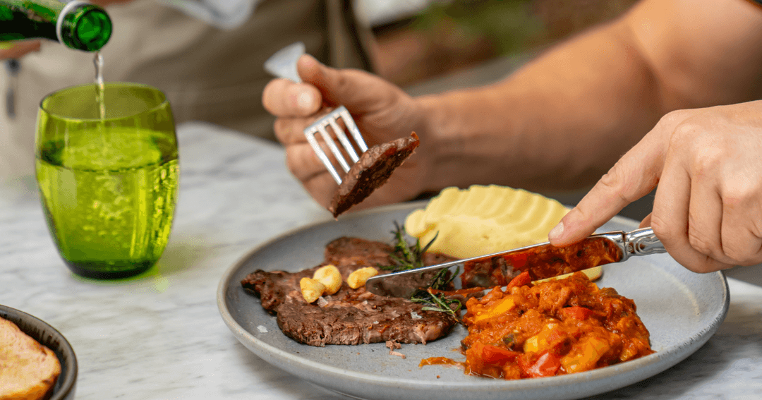  A person cuts into a steak on a plate with mashed potatoes and a vegetable stew, with a green glass of water beside it.