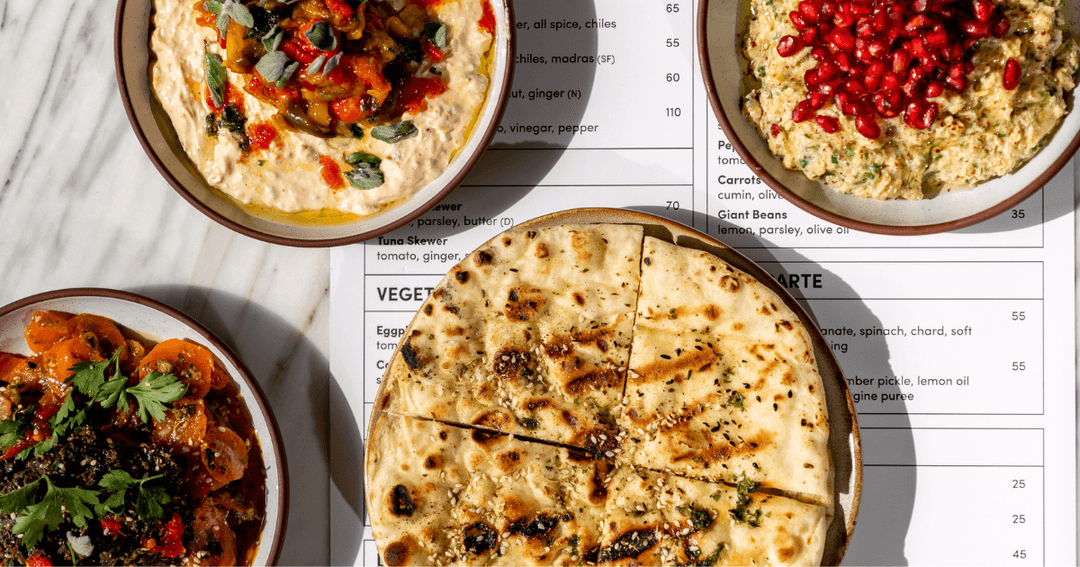 A variety of Mediterranean dishes displayed on a table, featuring hummus topped with herbs, a vegetable salad, and grilled flatbread, accompanied by a detailed menu.