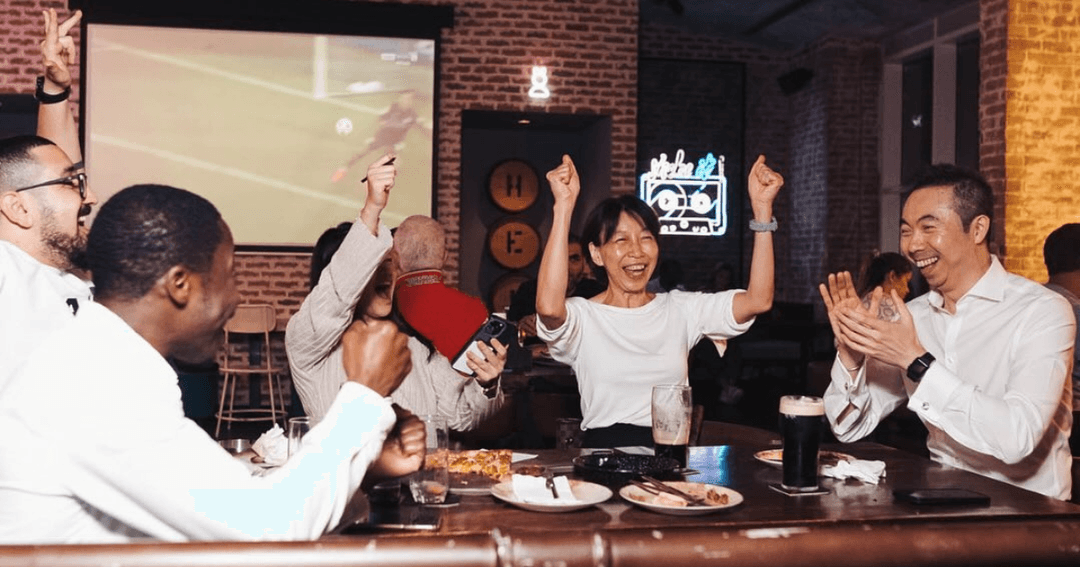 Joyful patrons at Brew House celebrate with drinks and cheers, enjoying a vibrant sports viewing experience under ambient lighting.