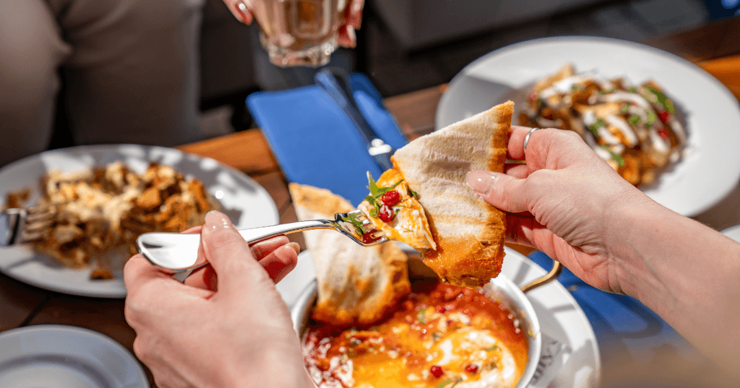 Hands holding bread scoop sauce from a breakfast dish, with other meals visible on the table.