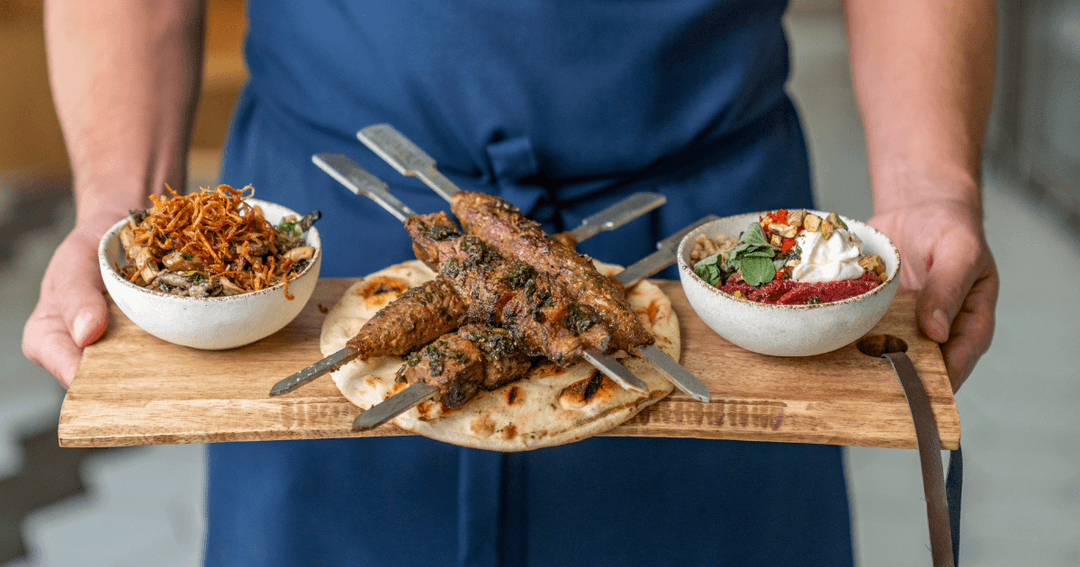 A server in Assembly restaurant, Expo City Dubai, presents a wooden platter with skewered kebabs, a bowl of mixed vegetables, and a dish of spiced condiments