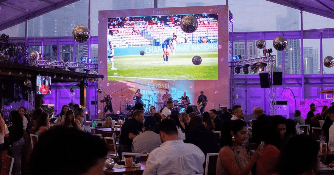 Live band performing on stage at Barasti Beach, with a large screen showing a sports event and a lively crowd enjoying the atmosphere.