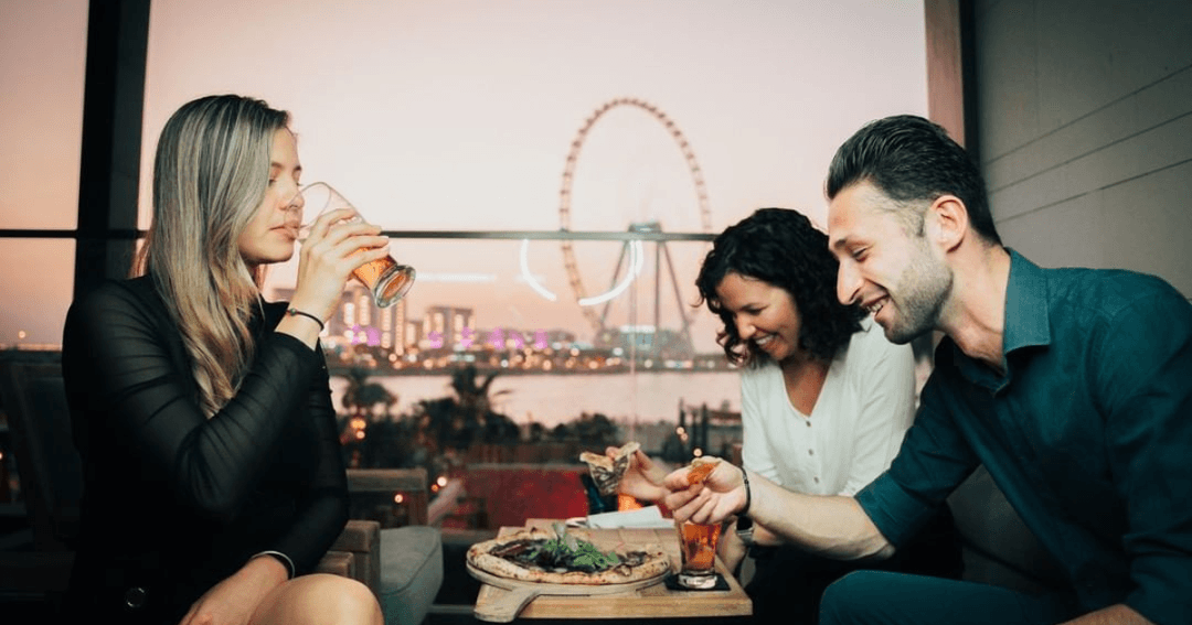 Three friends enjoying drinks and pizza with a stunning view of the Dubai Eye from the rooftop terrace at Bla Bla Dubai.