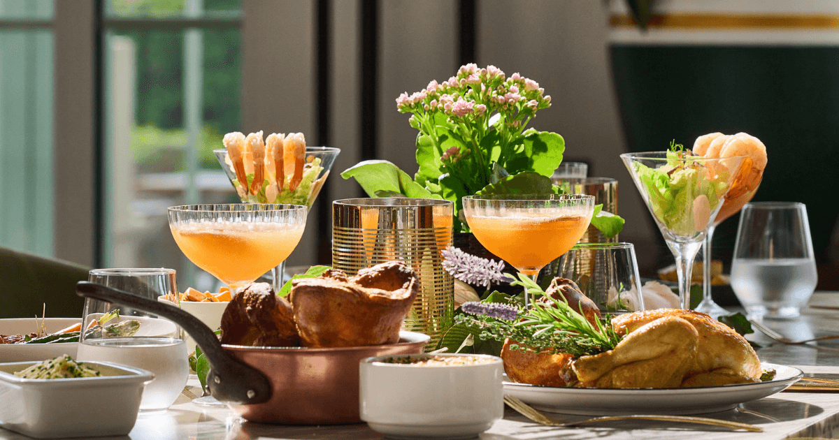 A beautifully arranged brunch table at Mina's Kitchen, featuring cocktails, shrimp cocktail appetizers, roasted chicken, and various side dishes, with vibrant greenery in the background.