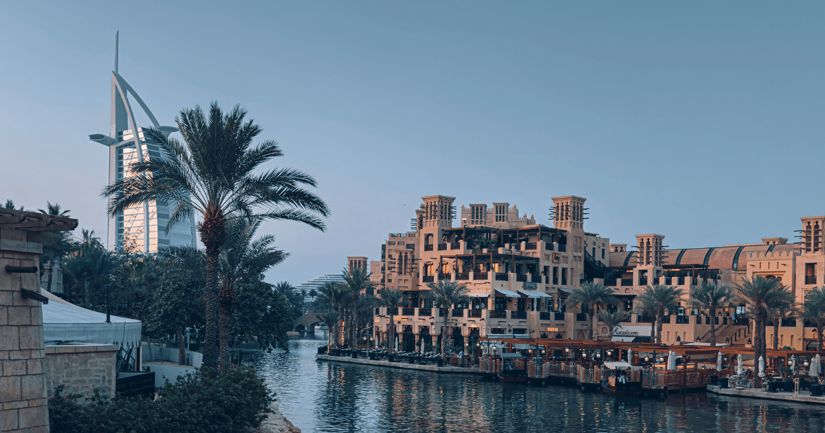 "A serene view of the Madinat Jumeirah complex in Dubai, showcasing traditional Arabian architecture alongside the tranquil waterways lined with palm trees. The iconic Burj Al Arab hotel stands prominently in the background against a clear evening sky."