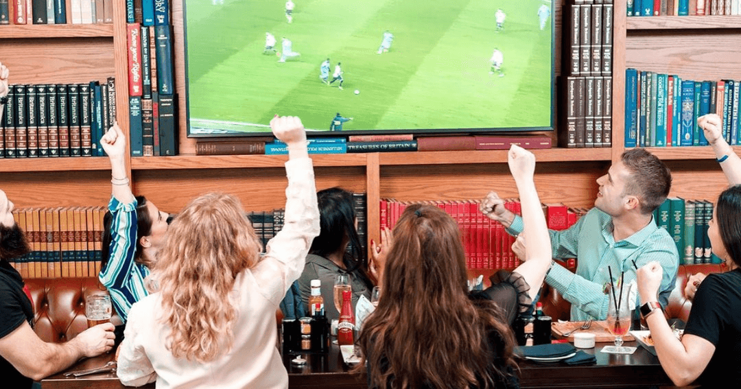 A group of friends cheering while watching a football match on a big screen at McGettigan's, Dubai, with a library-themed backdrop.