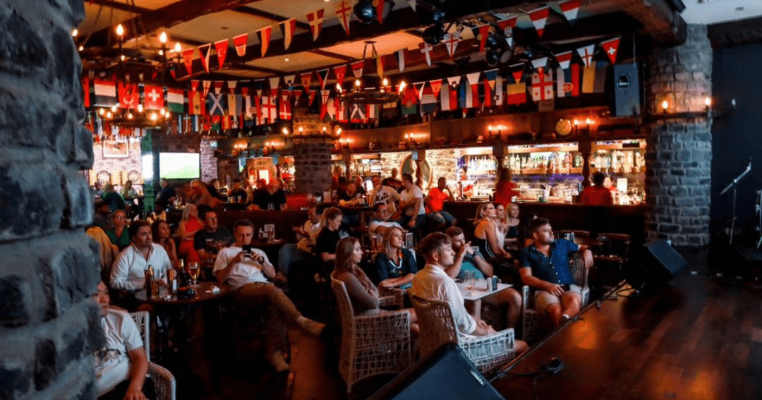 Crowd watching a sports event at Nell Gwynne, Dubai, with international flags decorating the pub and a lively atmosphere.